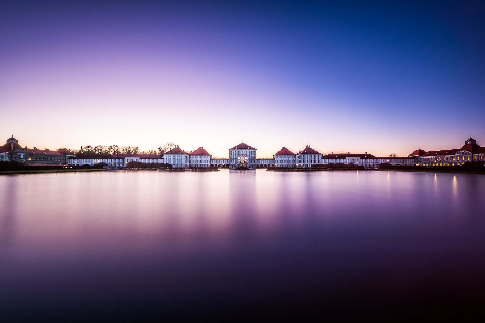 Nymphenburg Palace stretches across a still reflecting pool beneath a violet-blue evening sky.