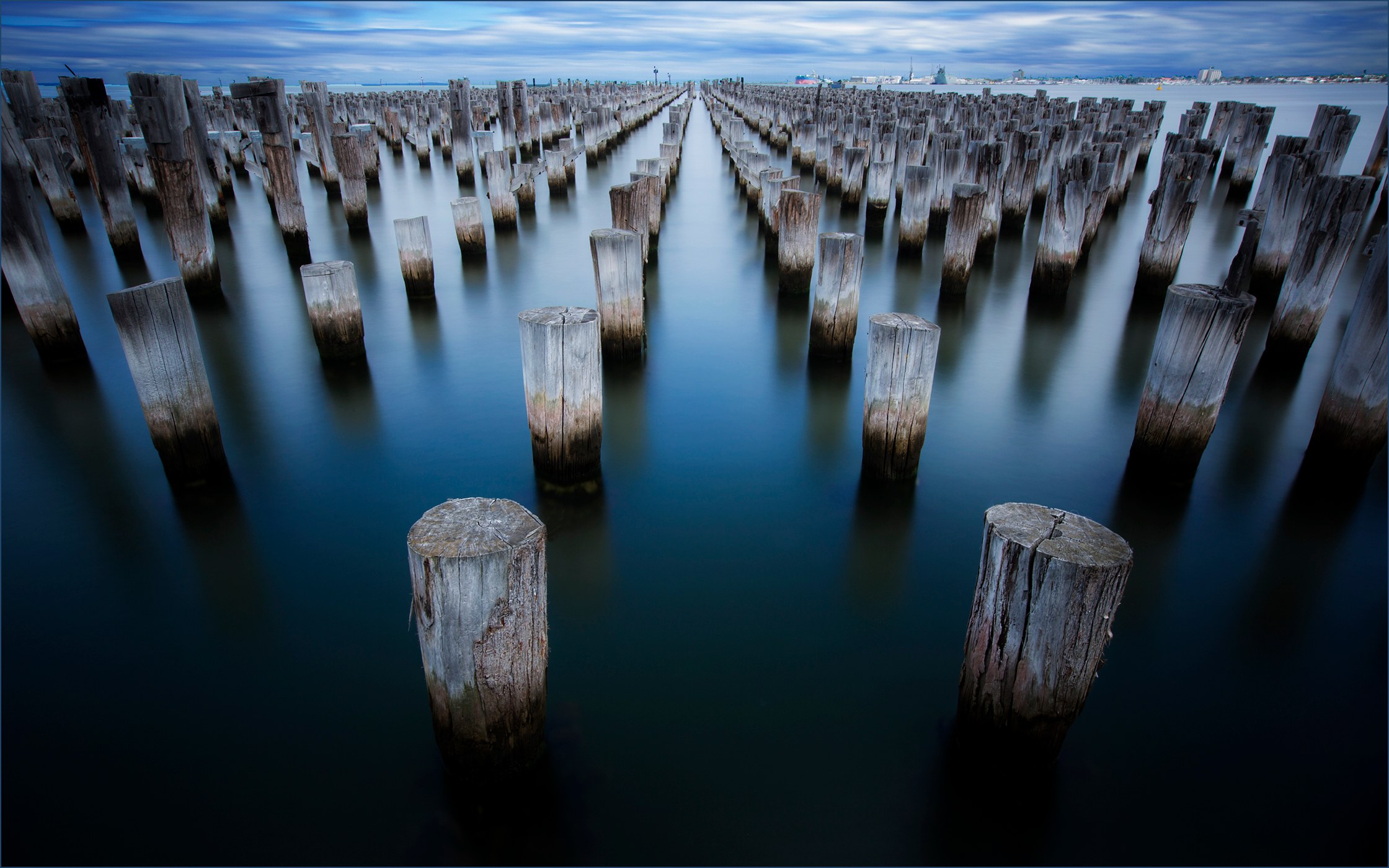 Rows of weathered wooden pilings recede through smooth blue water beneath streaked clouds.