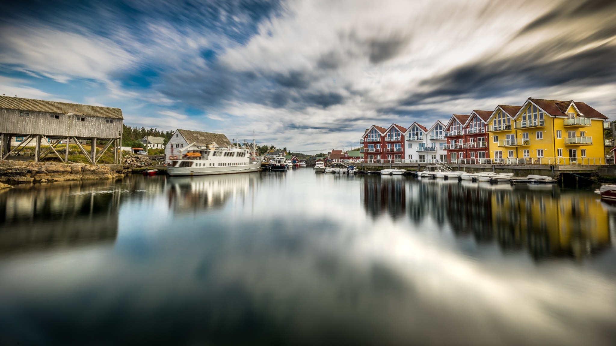 Colorful waterfront houses and a passing ferry reflect in a Norwegian harbor beneath fast-moving clouds.