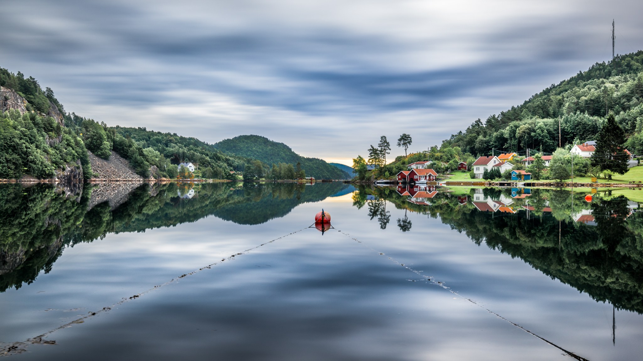 Red and white houses line a still Norwegian inlet, reflected between forested hills beneath streaked clouds.