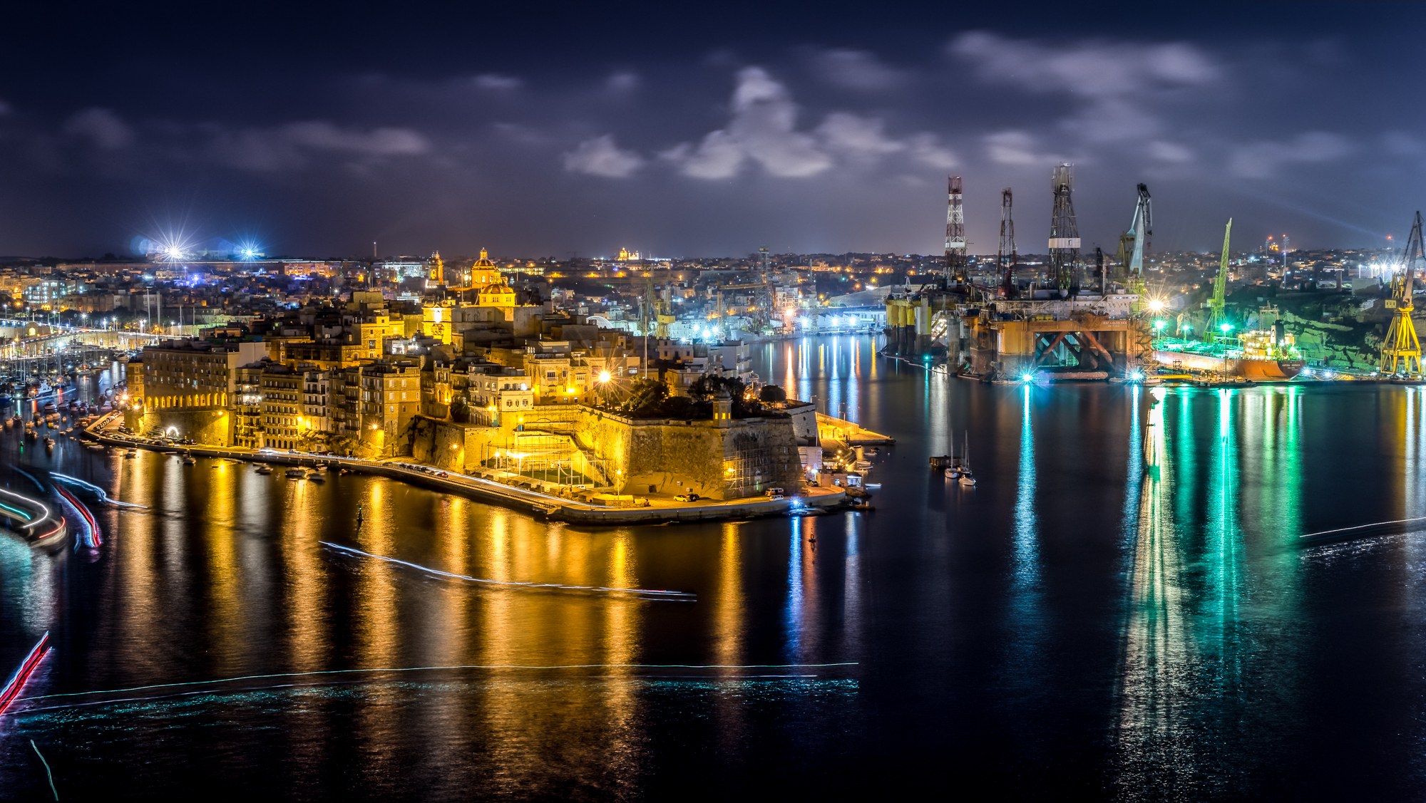 Golden waterfront buildings and working harbor cranes shine across dark water traced by passing boat lights.