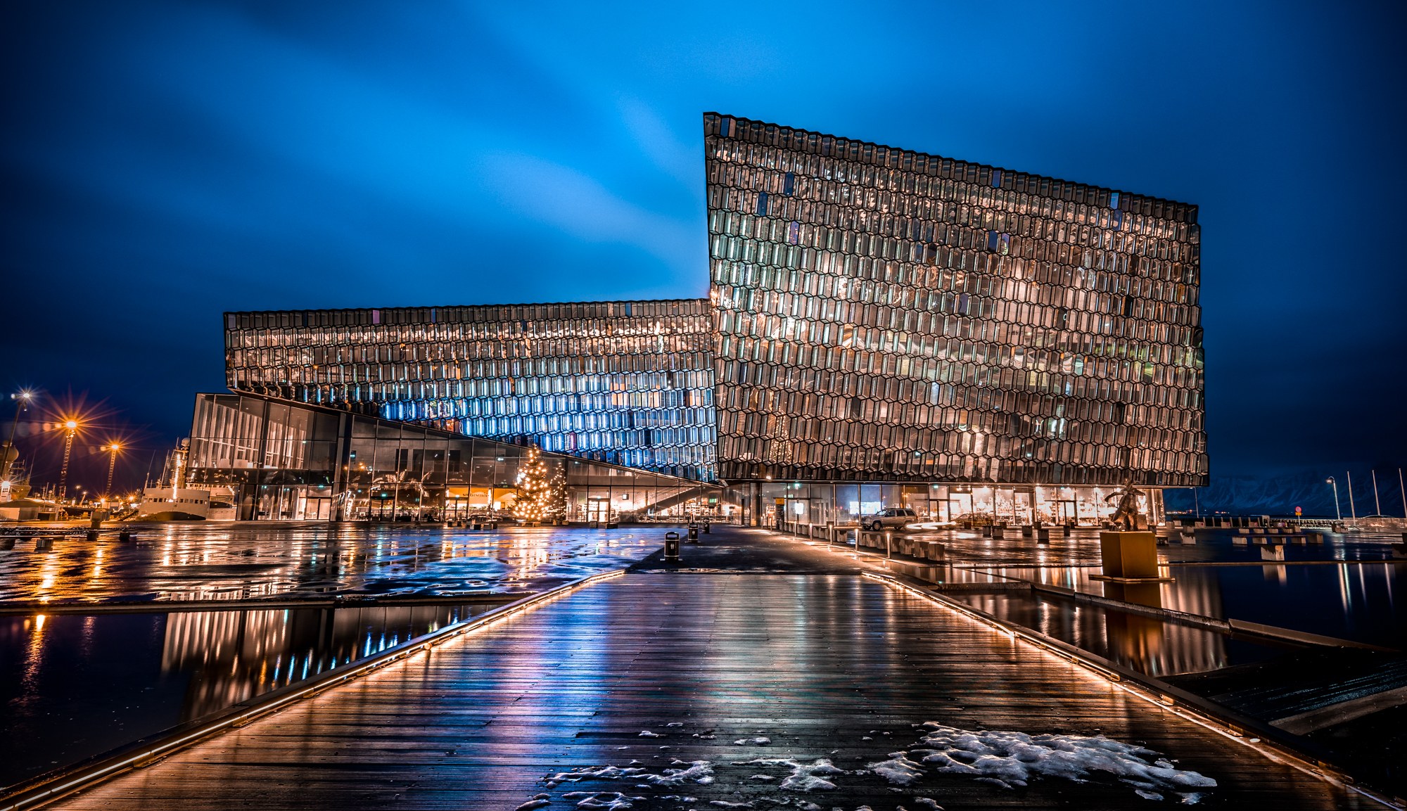 Harpa Concert Hall glows above a rain-slicked plaza and wooden walkway under a deep blue evening sky.