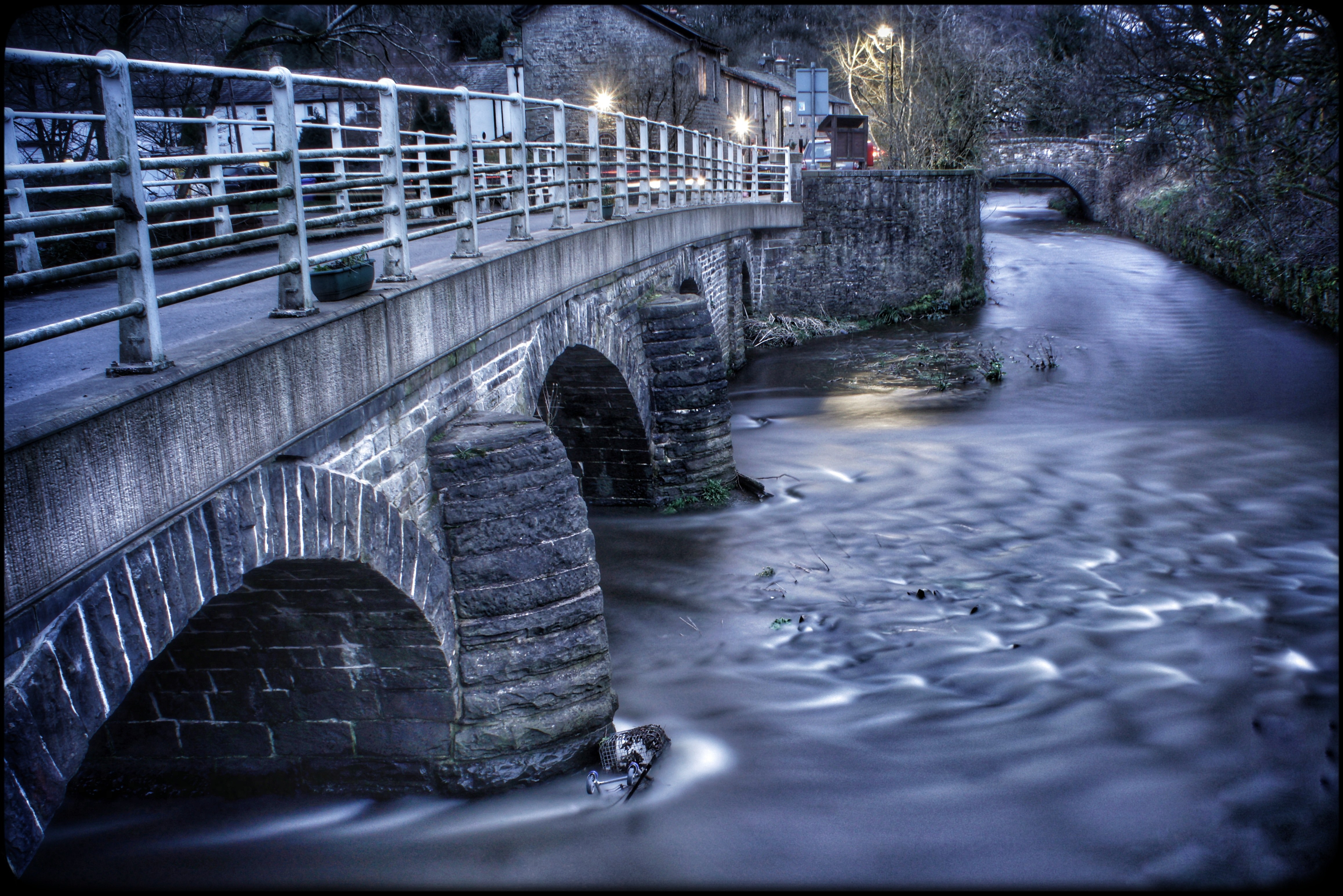 A metal-railed bridge crosses a smooth, fast-flowing river beside stone buildings and winter trees at blue hour.
