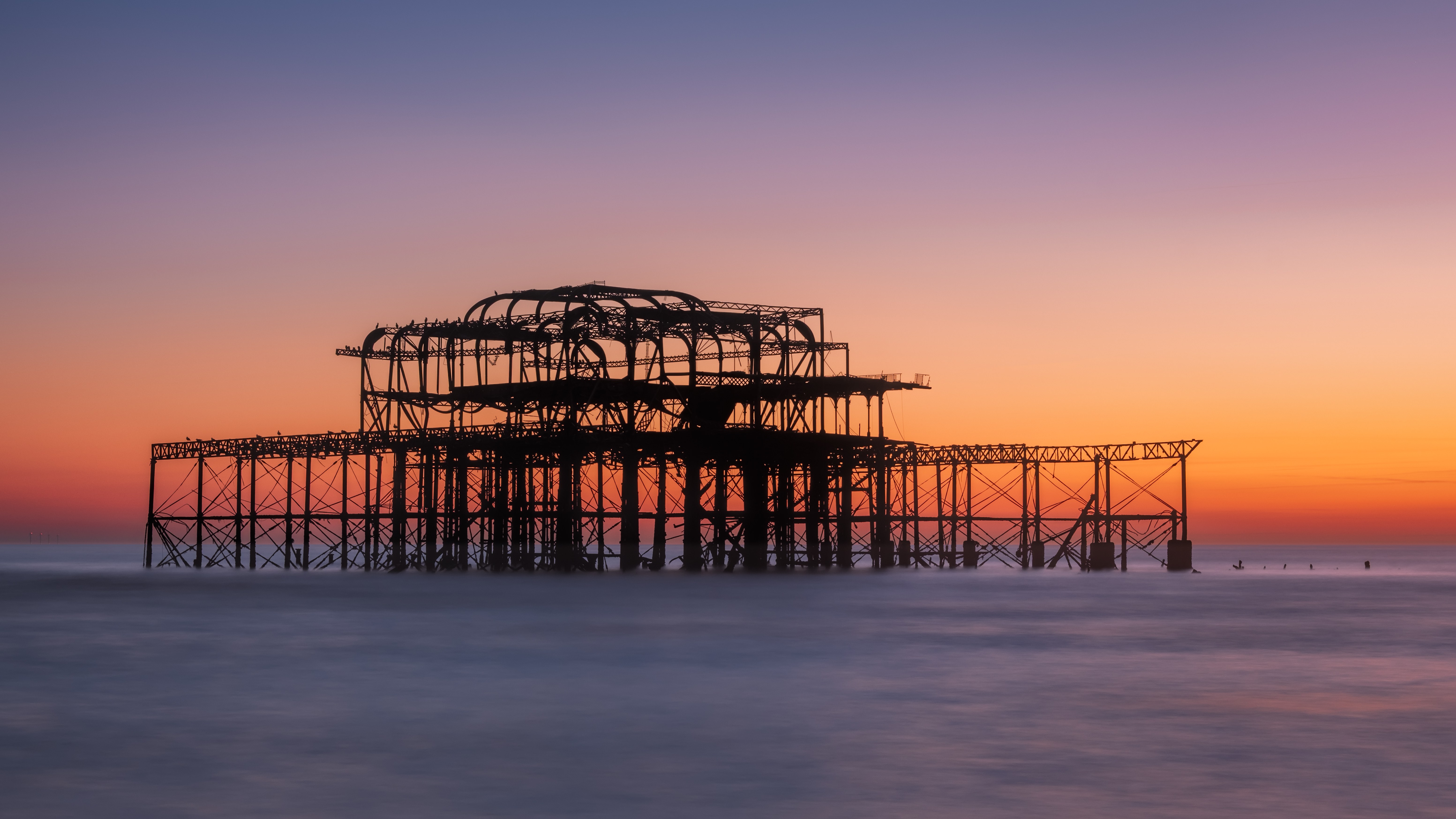 The skeletal remains of a seaside pier form a dark silhouette against a peach and lavender twilight sky above smooth water.
